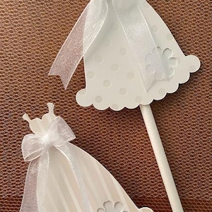 May include: Two white wooden dress-shaped ornaments with white ribbon bows and a white flower design. The ornaments are on sticks and are perfect for decorating a wedding or bridal shower.
