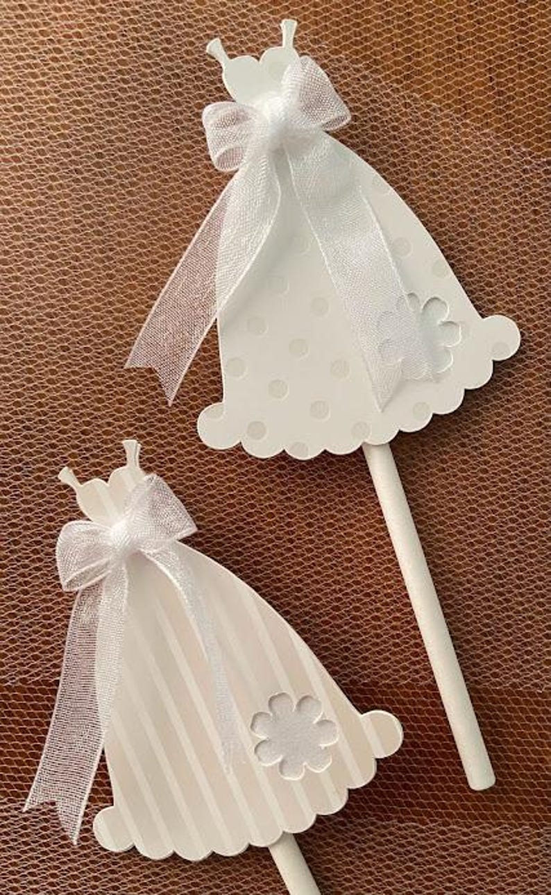 May include: Two white wooden dress-shaped ornaments with white ribbon bows and a white flower design. The ornaments are on sticks and are perfect for decorating a wedding or bridal shower.