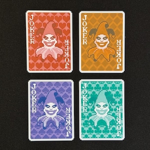 May include: Four playing cards featuring a pixelated joker design. Each card has a different background color: red, orange, purple, and green. The word "JOKER" is printed vertically on both sides of each card.