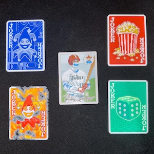 Puede incluir: Cinco cartas de juego con el tema "Joker". Las cartas presentan varios diseños, incluyendo un bufón, palomitas de maíz, un jugador de béisbol y un dado. Cada carta tiene la palabra "Joker" impresa.