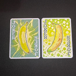 Puede incluir: Dos cartas de juego con un diseño de plátano pixelado. La palabra "JOKER" está impresa verticalmente en los lados izquierdo y derecho de cada carta. El fondo de una carta es verde y la otra es verde claro.