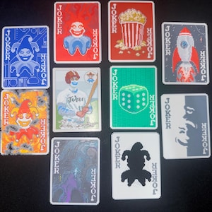 Pode incluir: Dez cartas de baralho com a palavra «JOKER» impressa. As cartas apresentam ilustrações pixelizadas: um palhaço, pipocas, um foguete, um jogador de beisebol, um dado e desenhos abstratos. Disposição em fundo escuro.