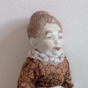 Geisha-granny collectible ceramic doll figurine, enigmatic blue-eyed face, floral lace dress and apron, vintage decor of ceramic and fabric