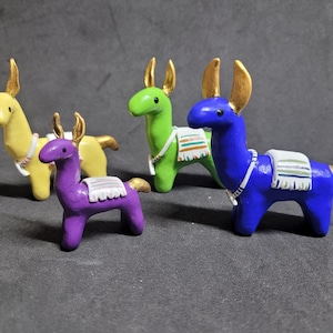 May include: Four colorful llama figurines. The llamas are painted in yellow, green, purple, and blue. Each has gold-colored ears and tail, and a decorative blanket. The llamas are standing on a gray surface.