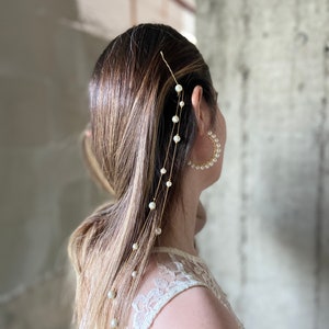 Può includere: Una catena per capelli dorata con perle bianche che pendono da un fermaglio dorato. La catena è indossata nei capelli di una donna, con il fermaglio posizionato vicino all'orecchio.