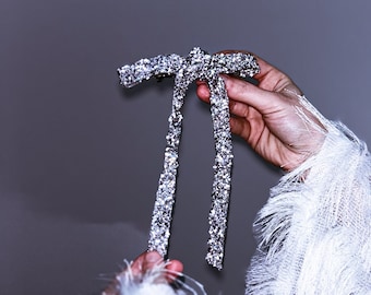 May include: A silver sparkly bow tie with two long tails. The bow tie is being held by a hand wearing a white fluffy sweater.