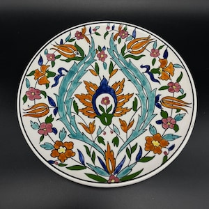 Może przedstawiać: Biały ceramiczny talerz z kolorowym wzorem kwiatowym. Wzór przedstawia pomarańczowe, niebieskie, zielone i różowe kwiaty i liście. Talerz jest tradycyjnym wzorem tureckim.