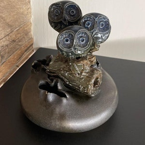 Könnte beinhalten: Eine Keramik-Eulen-Skulptur mit drei Eulen, die auf einem Ast sitzen. Die Eulen sind in Blautönen und Grün mit schwarzen Augen bemalt. Die Skulptur steht auf einem schwarzen Sockel.