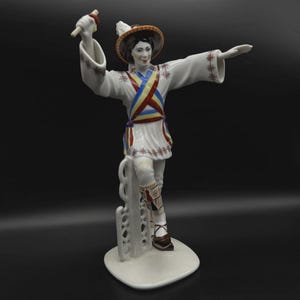 Puede incluir: Una figura de porcelana blanca de un bailarín con vestimenta tradicional. La figura lleva un sombrero, una túnica blanca con detalles rojos, azules y amarillos, y zapatos marrones. El bailarín posa con un brazo levantado y el otro extendido.