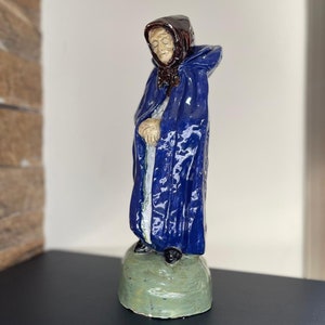 Op de afbeelding: Een keramiek beeldje van een oudere vrouw die een blauwe mantel en een bruine kap draagt. Ze staat op een groene basis.