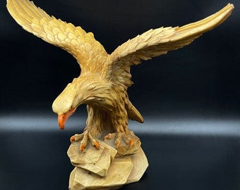 May include: A detailed sculpture of an eagle with outstretched wings, carved from a light tan material. The eagle's beak is orange, and its talons are a similar color. The sculpture is mounted on a rock-like base, showcasing intricate details.
