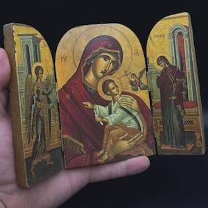 Pode incluir: Ícone tríptico de madeira com figuras religiosas. O painel central mostra uma mulher segurando uma criança, cercados por halos. Os painéis laterais mostram outras figuras com roupas detalhadas e coloridas. O ícone é segurado em uma mão.