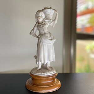 Puede incluir: Una figurita de porcelana blanca de una niña que lleva una cesta de fruta. La figurita está sobre una base de madera.