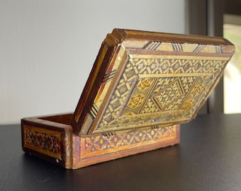 May include: A small wooden box with a hinged lid. The box is decorated with intricate geometric patterns in shades of brown, gold, and white. The lid is open, revealing the interior of the box.