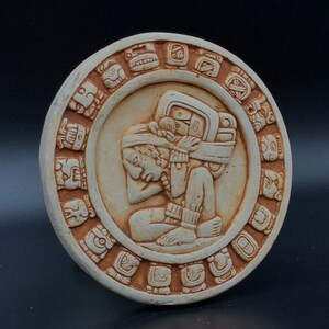 May include: A beige, circular relief sculpture depicting a Mayan figure carrying a burden, surrounded by glyphs. The figure is in a seated position, with detailed carvings. The outer ring features a series of carved symbols. The sculpture is set against a dark background.
