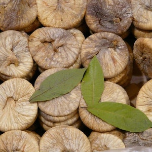 May include: A close-up of dried figs, some stacked on top of each other, with two green leaves on top. The figs are a light brown colour and have a wrinkled texture.