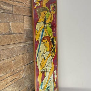 May include: A vertical wooden artwork featuring a painted illustration of three figures in white dresses. The artwork has a red and yellow background with text in the upper left and right corners. The artwork is displayed against a brick wall.