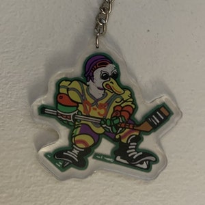 May include: A clear plastic keyring featuring a cartoon duck in a hockey uniform, holding a hockey stick. The duck is wearing a yellow jersey with purple and green accents, a purple helmet, and black skates. The keyring has a silver ring and chain.