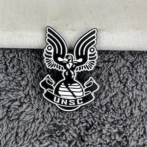 May include: Black and white enamel pin featuring the UNSC logo from the Halo video game series. The logo depicts a stylised eagle with wings spread, holding a sphere with the letters UNSC.