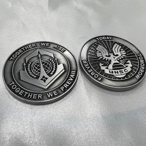 May include: Two silver-colored metal coins with different designs. The first coin features a stylized logo with the text "Together We Rise Together We Prevail". The second coin features a stylized eagle logo with the text "Today Forever Tomorrow" and "UNSC".