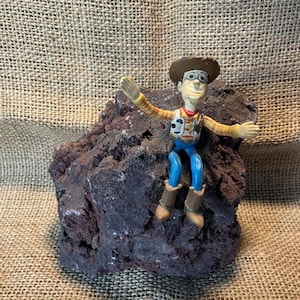 May include: A small plastic toy figurine of Woody from Toy Story sitting on a dark gray rock. Woody is wearing his signature cowboy hat, vest, and blue jeans.