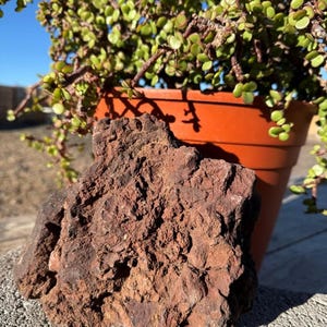 May include: A rough, porous, reddish-brown volcanic rock with an irregular, craggy surface texture. The stone features deep crevices and a matte finish, displaying a natural, earthy appearance with varied dark and light brown color tones throughout.