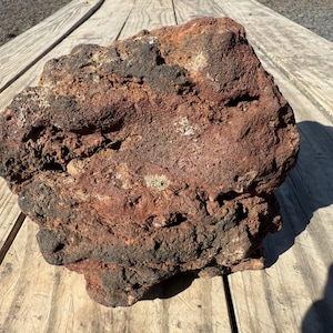 May include: A large, irregularly shaped rock with a rough, porous texture. The rock's color is primarily a deep reddish-brown, with patches of black and lighter tan hues. The rock is sitting on a wooden surface.