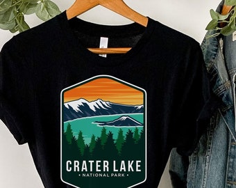 May include: Black t-shirt with a vintage-style graphic of Crater Lake National Park. The graphic features a mountain range, a lake, and trees in shades of green, blue, and orange.