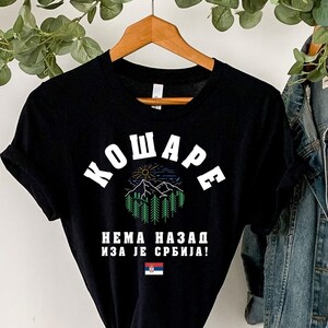 Puede incluir: Camiseta negra con texto blanco que dice "KOWARE NEMA NAZAD IZA JE SRBIJA!" con un gráfico de una cordillera y un sol. La camiseta está colgada en una percha con una chaqueta vaquera.