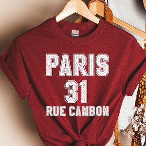 Pu&ograve; includere: Una maglietta color granata con testo bianco che recita "PARIS 31 RUE CAMBON".