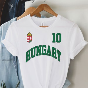 May include: White t-shirt with the word "HUNGARY" in green block letters. The number "10" is above the word, also in green. A Hungarian crest is on the upper left. The shirt is hanging on a wooden hanger.