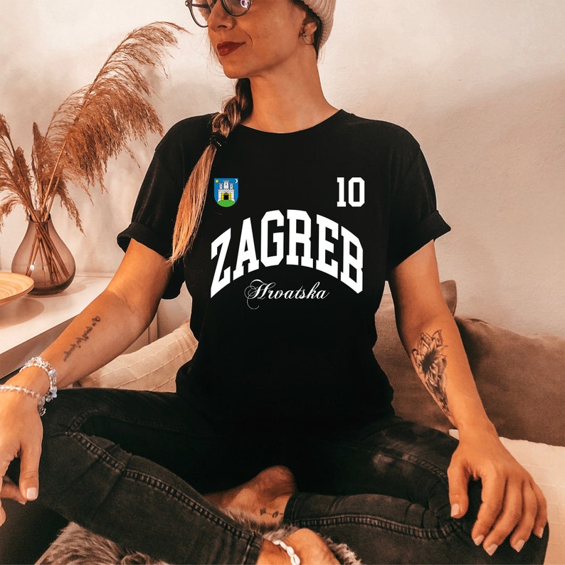 Puede incluir: Camiseta negra con la palabra "ZAGREB" en blanco, el n&uacute;mero "10" y la palabra "Hrvatska" en cursiva. Un peque&ntilde;o emblema est&aacute; en la parte superior izquierda. La camiseta la lleva una persona.