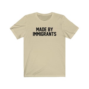 May include: A beige t-shirt with the text "MADE BY IMMIGRANTS" printed in black.