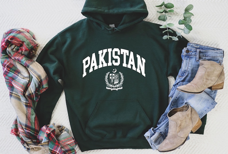 May include: A dark green hooded sweatshirt with the word "PAKISTAN" in white arched lettering. Below the word is a white emblem. The sweatshirt is paired with blue jeans, tan boots, and a plaid scarf.