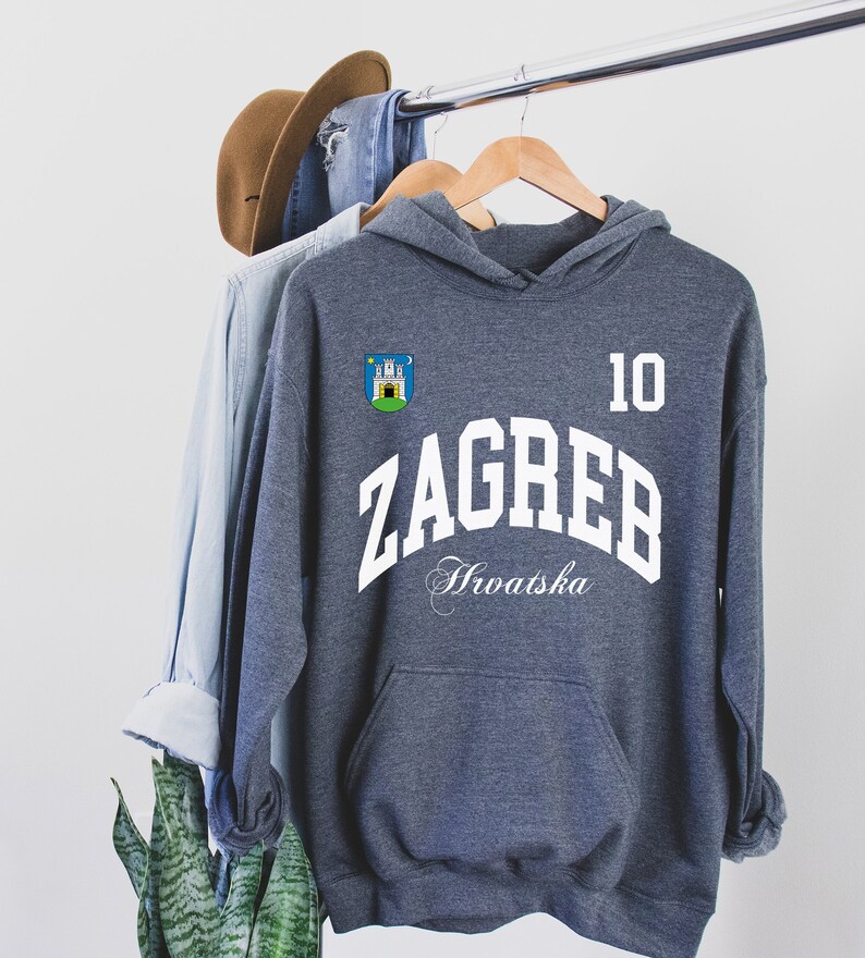 May include: A gray hoodie with the word "ZAGREB" in white block letters, and the number "10". The hoodie also features a crest and the word "Hrvatska" in script. A brown hat and denim shirt hang on the rack behind the hoodie.