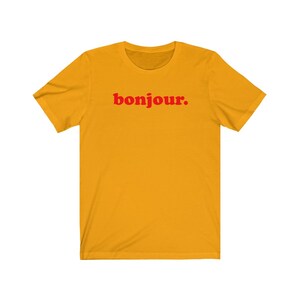 Pode incluir: Uma camiseta amarela de manga curta com a palavra "bonjour." impressa em vermelho.