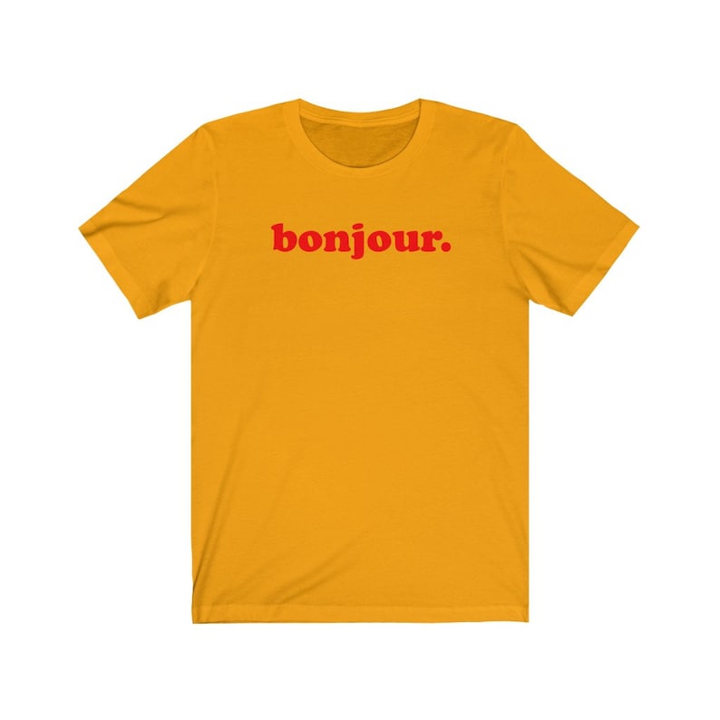 Pode incluir: Uma camiseta amarela de manga curta com a palavra "bonjour." impressa em vermelho.