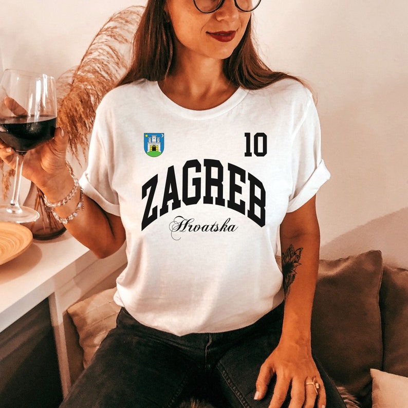 May include: White t-shirt with the word "ZAGREB" in black block letters, the number "10", and the word "Hrvatska" in script. A small emblem is above the word "ZAGREB". The shirt is worn by a person.