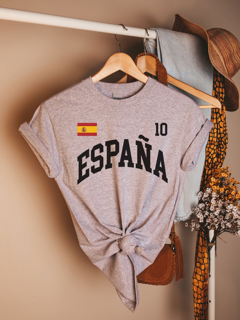 K&ouml;nnte beinhalten: Ein meliertes graues T-Shirt mit dem Wort "ESPA&Ntilde;A" in schwarzer, gebogener Schrift. Die spanische Flagge befindet sich oben links und die Nummer "10" oben rechts. Das T-Shirt ist an der Taille geknotet.