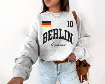 May include: A light gray sweatshirt with the word "BERLIN" in bold black letters, the number "10", and the German flag. Below "BERLIN" is the word "Germany" in a script font. The sweatshirt is a casual style.