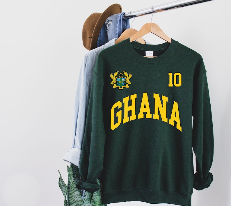 May include: A dark green sweatshirt with the word "GHANA" in yellow arched lettering. The number "10" is printed in yellow on the upper right, and a crest is on the upper left. A brown hat and blue shirt hang on the rack behind the sweatshirt.