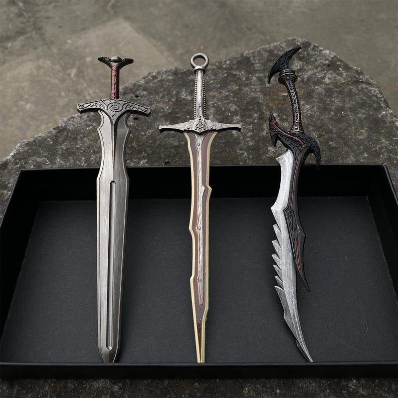 May include: Three decorative swords with intricate designs. The swords feature silver, brown, and black blades and handles. The swords are displayed in a black box on a dark gray surface. The swords have unique hilt and blade shapes.