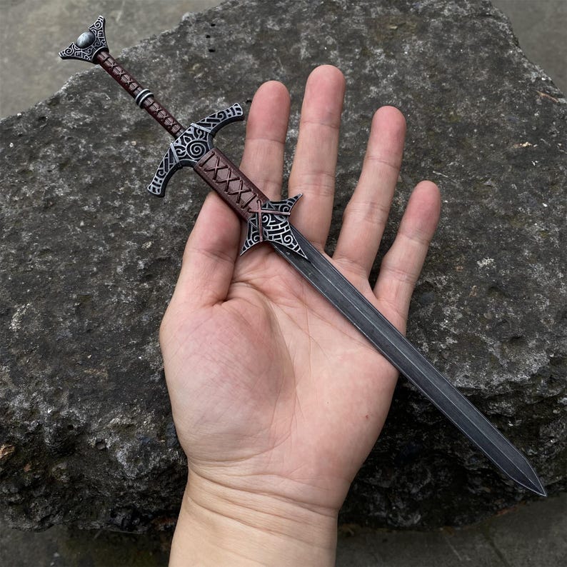 May include: A miniature sword with a dark gray blade and ornate silver-colored hilt. The handle is wrapped in brown leather. The sword is held in a hand against a textured gray stone background. The sword measures approximately 10 inches in length.