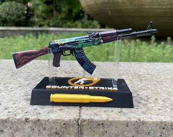 May include: A miniature rifle model featuring a wood-grain stock and a green and black graphic pattern on the barrel. It is mounted on a black display stand with the text "COUNTER-STRIKE" and a gold-colored bullet replica attached.