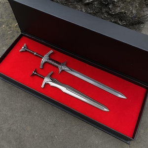 May include: Two decorative swords in a black presentation box with a red velvet lining. The swords have silver blades and ornate handles. The larger sword is approximately 24 inches long, and the smaller sword is about 10 inches long.