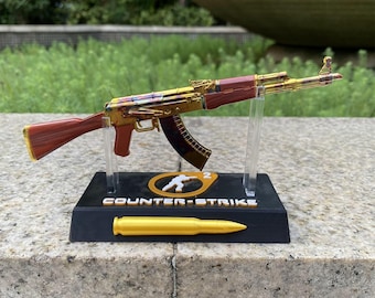 May include: A miniature gold and reddish-brown rifle model mounted on a black display stand. The base features the text COUNTER-STRIKE 2 and a gold bullet replica. The rifle is supported by two clear acrylic pillars on the black rectangular base.