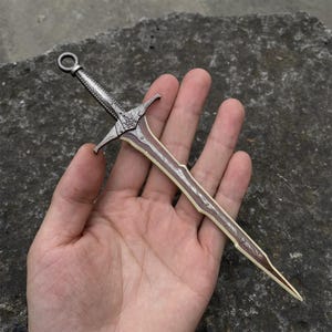 May include: A miniature decorative sword with a silver-colored hilt and a blade featuring brown and cream accents. The sword has a detailed design and a ring at the top of the hilt.