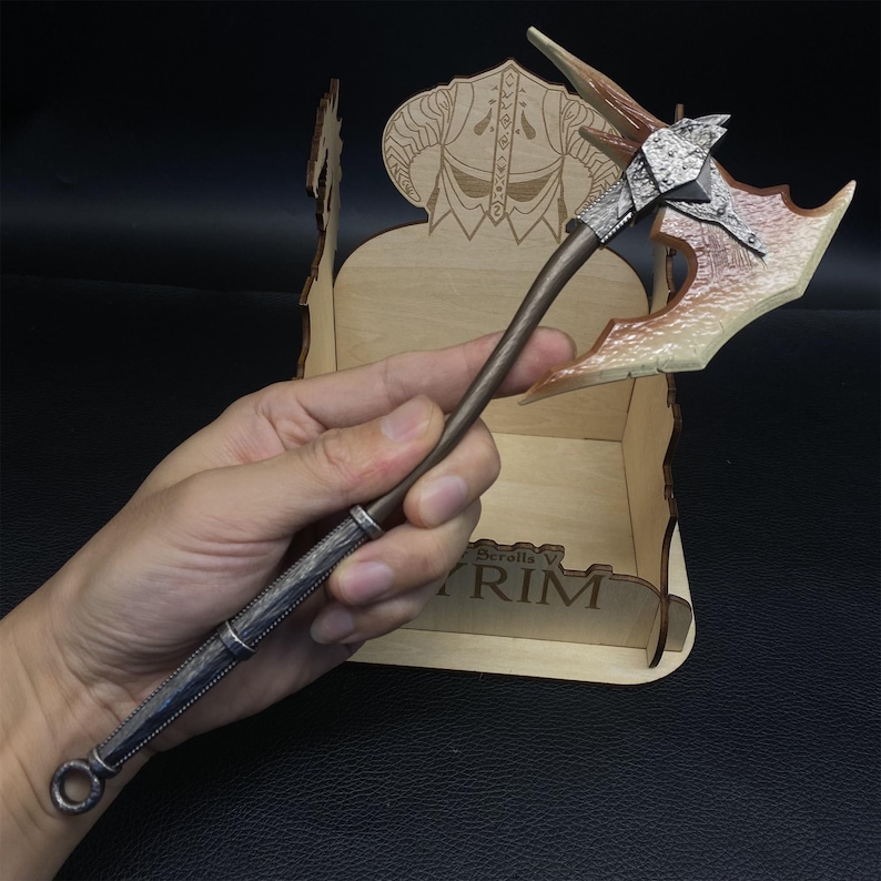 May include: A miniature axe with a brown handle and a silver-colored head. The axe head has a unique design with a light brown and white color scheme. The axe is held in front of a wooden display stand with the word "SKYRIM" visible.