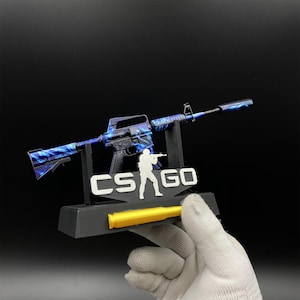Pu&ograve; includere: Replica in miniatura di un fucile con un motivo a vortice blu e nero. Il fucile &egrave; montato su un supporto nero con la scritta "CS GO" e la silhouette di una persona che tiene un'arma. Un proiettile giallo &egrave; posto alla base.