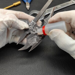 May include: A person wearing white gloves is applying clear glue to a metal object with a detailed design. The object has a sharp, pointed shape and appears to be a decorative piece.